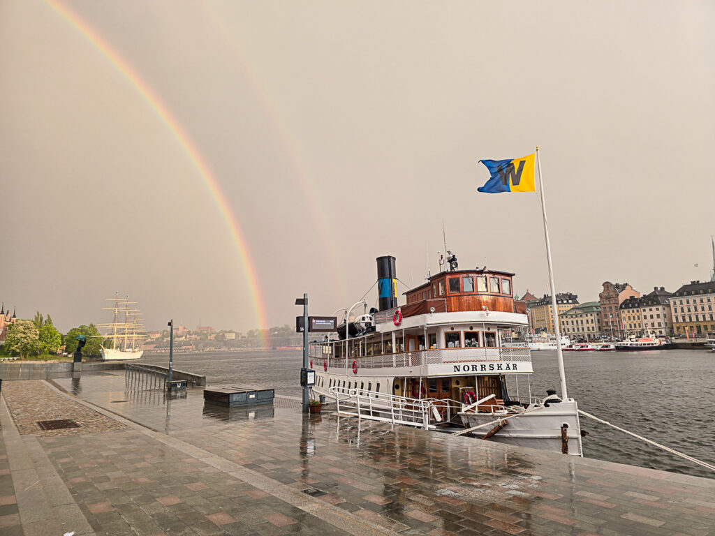 Regnbåge över Stockholm och skärgårdsbåt vid Skeppsholmen